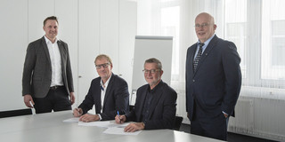 Dr. Ronald Kriedel und Prof. Andreas Liening stehen an einem Tisch. Am Tisch sitzen Albrecht Ehlers und Guido Baranowski und unterzeichnen Verträge