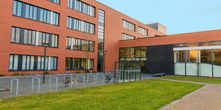 Seminarraumgebäude: Ein rotes dreistöckiges Gebäude mit schwarz gerahmten Fenstern, von der Seite fotografiert. Davor eine Wiese und Fahrradständer sowie Straßenlaternen.