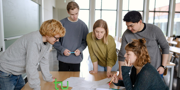 Studierende Gruppenarbeit Fünf Studierende stehen um einen Tisch und sehen auf ein Dokument.