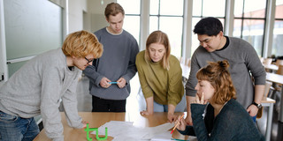 Students group work_02 Five students stand around a table and look at a piece of paper.