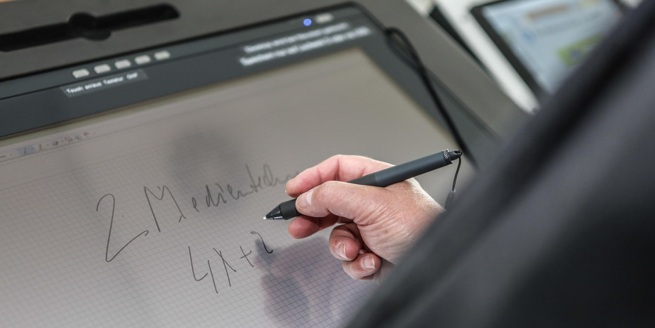 Bildschirm eines Hörsaalpults, an dem jemand mit einem Stift in der Hand schreibt.