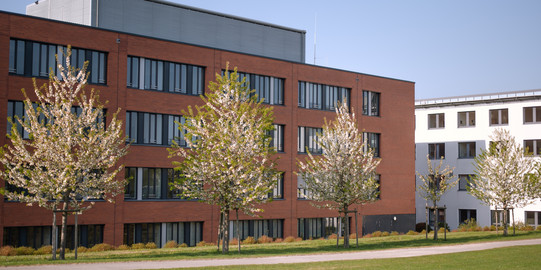 Ein rotes Backsteingebäude der TU Dortmund mit blühenden Bäumen und grüner Wiese im Sommer.
